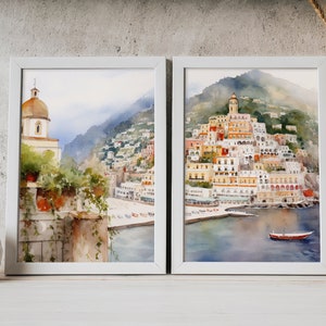 May include: Two framed watercolor paintings depicting a coastal Italian town with colorful buildings, a church, and a boat in the water. The paintings are set against a white wall with a vase of wheat stalks and a white ceramic vase.