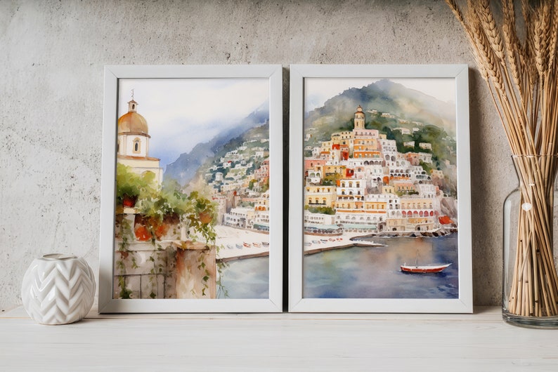 May include: Two framed watercolor paintings depicting a coastal Italian town with colorful buildings, a church, and a boat in the water. The paintings are set against a white wall with a vase of wheat stalks and a white ceramic vase.