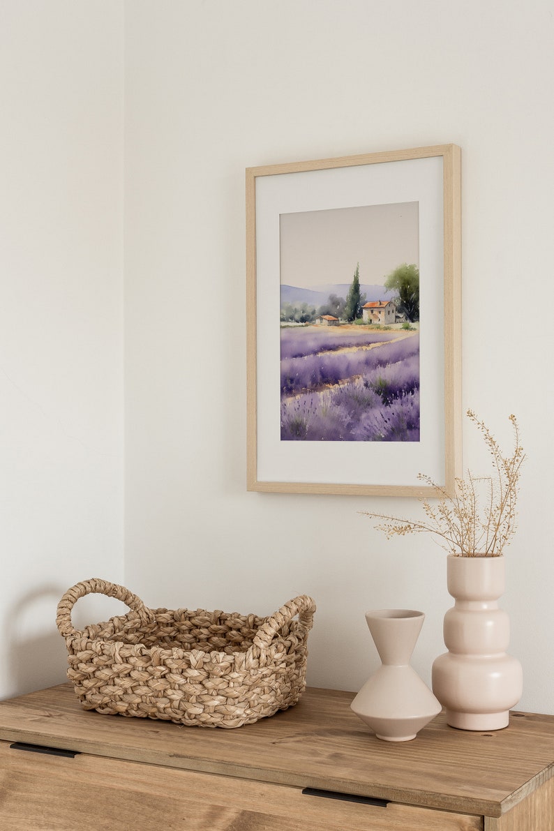 May include: A framed watercolor painting of a lavender field with a small house in the distance. The painting is hanging on a white wall above a wooden dresser with a woven basket and two ceramic vases.
