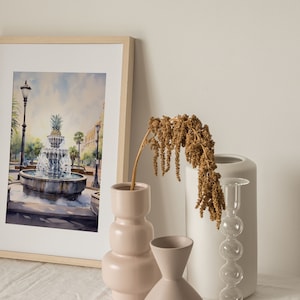 May include: A framed watercolor painting of a fountain with a pineapple on top, surrounded by palm trees and a street lamp. The painting is on a table with a vase of dried flowers, a white vase, and a clear glass vase with a bulbous shape.