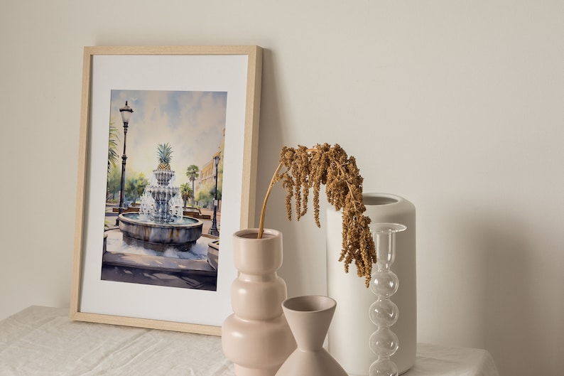 May include: A framed watercolor painting of a fountain with a pineapple on top, surrounded by palm trees and a street lamp. The painting is on a table with a vase of dried flowers, a white vase, and a clear glass vase with a bulbous shape.