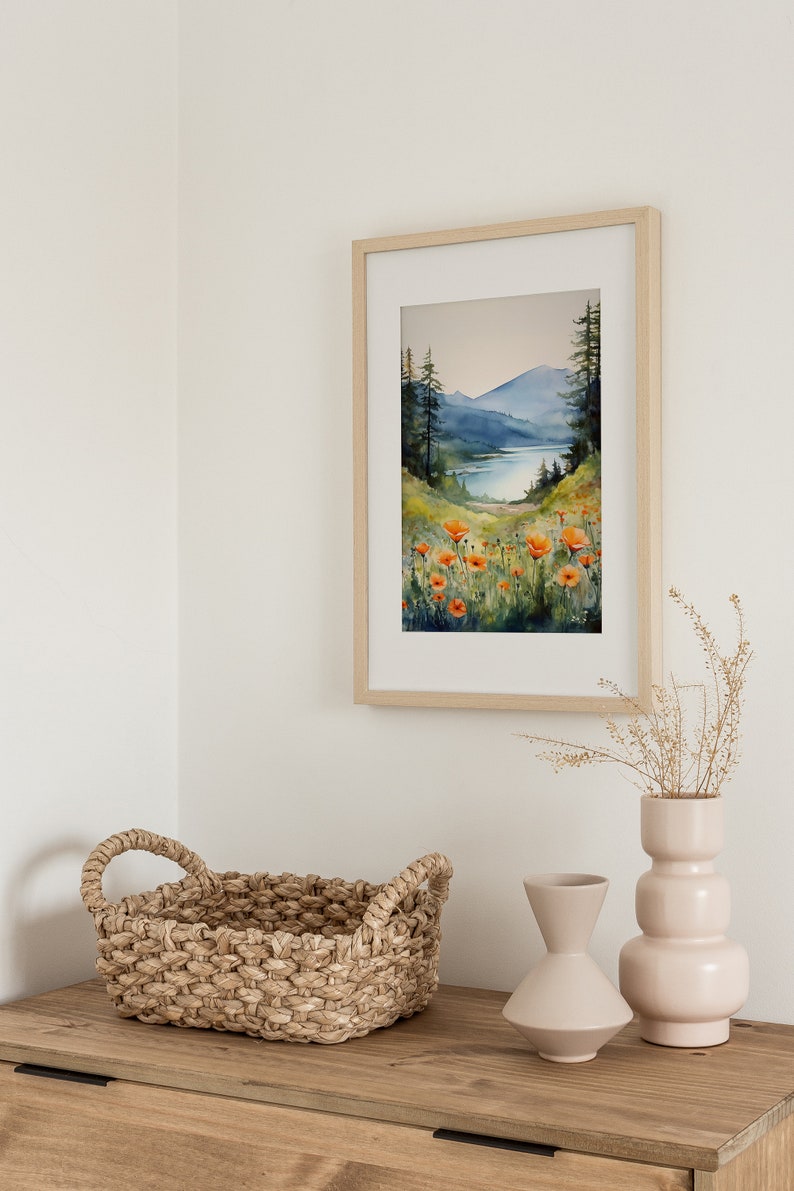 Puede incluir: Una pintura al agua enmarcada de una escena de lago de monta&ntilde;a con flores naranjas en primer plano. La pintura est&aacute; colgada en una pared blanca sobre una c&oacute;moda de madera con una cesta tejida y dos jarrones de cer&aacute;mica.