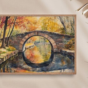 May include: Watercolor painting of an arched stone bridge over a calm river, framed in a wooden border. The artwork depicts an autumn scene with trees in shades of yellow, orange, and red reflected in the water. The bridge is made of stone.