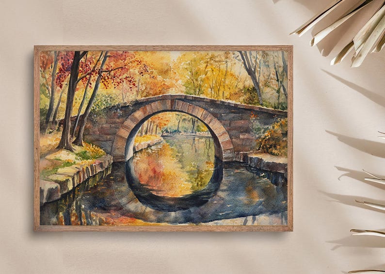 May include: Watercolor painting of an arched stone bridge over a calm river, framed in a wooden border. The artwork depicts an autumn scene with trees in shades of yellow, orange, and red reflected in the water. The bridge is made of stone.