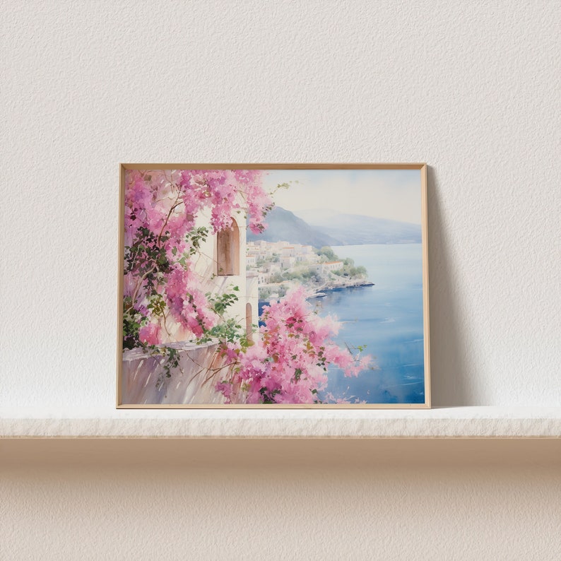 以下が含まれることがあります： 青い海と小さな町を見下ろす崖に生えているピンクの花の茂みを水彩で描いた絵画。絵画は明るい木製の額縁に入っています。