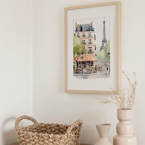 May include: A framed watercolor print of a Parisian street scene with a cafe and the Eiffel Tower. A woven basket and two light pink vases sit on a wooden surface. The artwork is in a light wood frame.