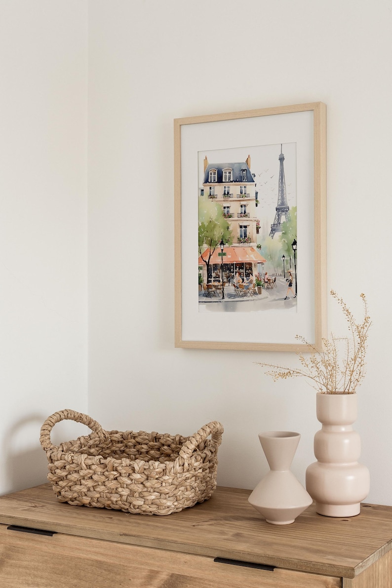May include: A framed watercolor print of a Parisian street scene with a cafe and the Eiffel Tower. A woven basket and two light pink vases sit on a wooden surface. The artwork is in a light wood frame.
