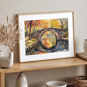 May include: Watercolor painting of a stone bridge over a calm river, framed in a light wood frame. The artwork depicts an autumn scene with vibrant orange and yellow foliage reflected in the water. The bridge has a circular arch.