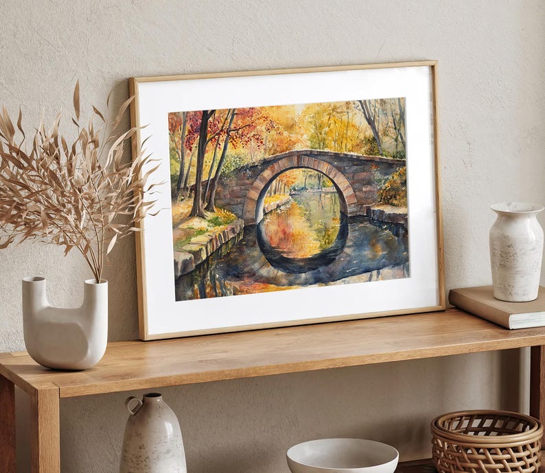 May include: Watercolor painting of a stone bridge over a calm river, framed in a light wood frame. The artwork depicts an autumn scene with vibrant orange and yellow foliage reflected in the water. The bridge has a circular arch.