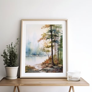 May include: A watercolor painting of a forest scene with a lake in the foreground. The trees are a mix of green and brown, and the water is a light blue. The painting is framed in a light wood frame.