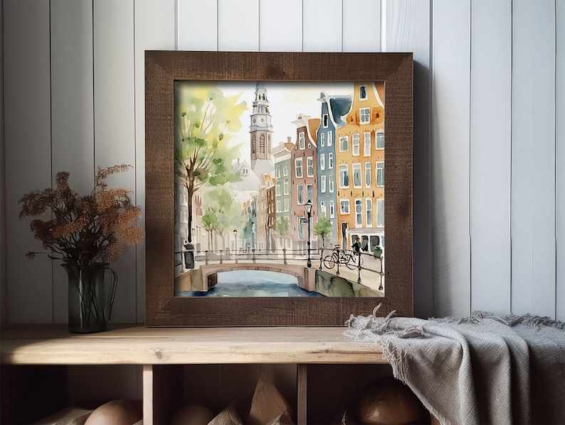 May include: A watercolor painting of a canal scene in Amsterdam, Netherlands. The painting features colorful buildings, a bridge, a canal, and a bicycle. The painting is framed in a brown wooden frame.
