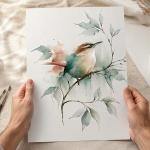 Könnte beinhalten: Ein Aquarellbild auf weißem Papier, das einen kleinen braunen und cremefarbenen Vogel auf einem dünnen Zweig zeigt. Der Vogel ist von weichen, gedämpften grünen Blättern und subtilen Pfirsich- und Blaugrüntönen in einem zarten Stil umgeben.