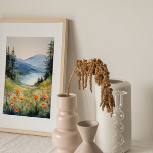 Puede incluir: Una pintura de acuarela enmarcada que representa una escena de lago de monta&ntilde;a con amapolas naranjas en primer plano. La pintura se exhibe en una mesa blanca con tres jarrones, uno con flores secas, uno blanco y uno rosa.