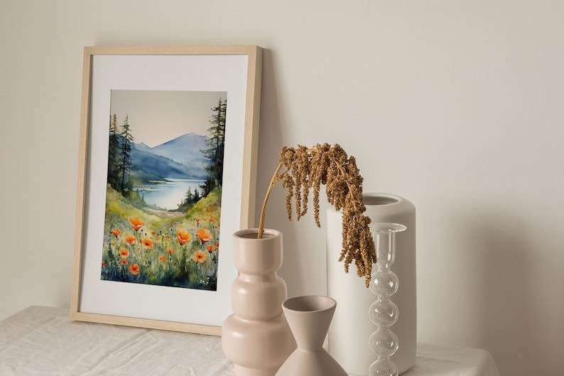 Puede incluir: Una pintura de acuarela enmarcada que representa una escena de lago de monta&ntilde;a con amapolas naranjas en primer plano. La pintura se exhibe en una mesa blanca con tres jarrones, uno con flores secas, uno blanco y uno rosa.