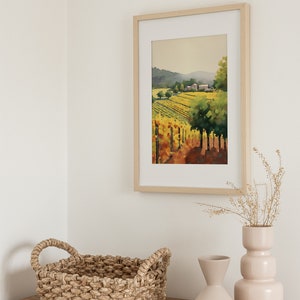 Puede incluir: Una pintura de acuarela enmarcada de un paisaje de vi&ntilde;edo con una granja en la distancia. La pintura est&aacute; en un marco de madera claro y est&aacute; colgada en una pared blanca. Hay dos jarrones con flores secas y una cesta tejida sobre una mesa de madera en primer plano.