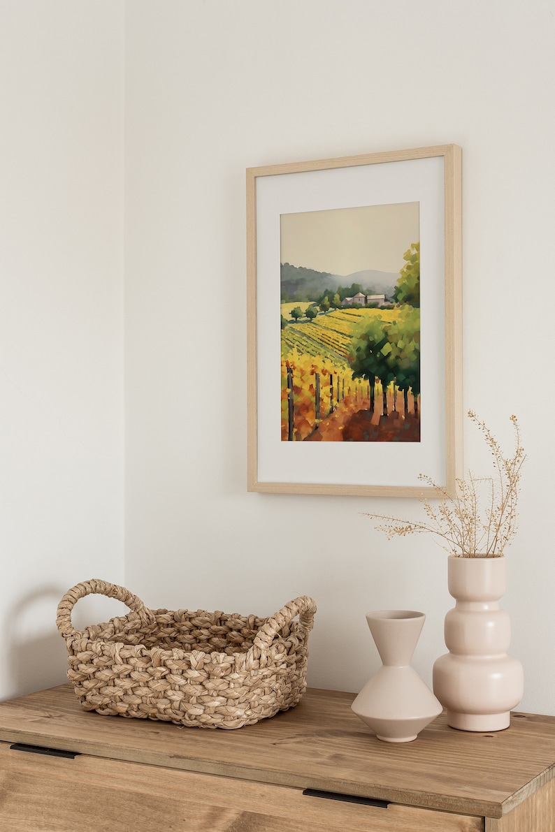 Puede incluir: Una pintura de acuarela enmarcada de un paisaje de vi&ntilde;edo con una granja en la distancia. La pintura est&aacute; en un marco de madera claro y est&aacute; colgada en una pared blanca. Hay dos jarrones con flores secas y una cesta tejida sobre una mesa de madera en primer plano.