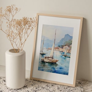 May include: A framed watercolour painting of a sailboat on a body of water with mountains in the background. The painting is in a light wood frame and is sitting on a white and black rug.