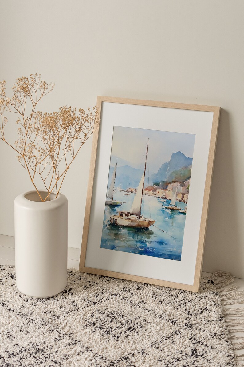 May include: A framed watercolour painting of a sailboat on a body of water with mountains in the background. The painting is in a light wood frame and is sitting on a white and black rug.