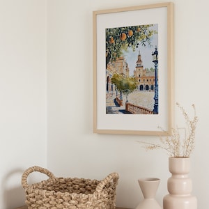 May include: A framed watercolour painting of a Spanish plaza with a church in the background, a tree with orange fruit, and a street lamp. The painting is hanging on a white wall above a wooden dresser with a woven basket and two ceramic vases.