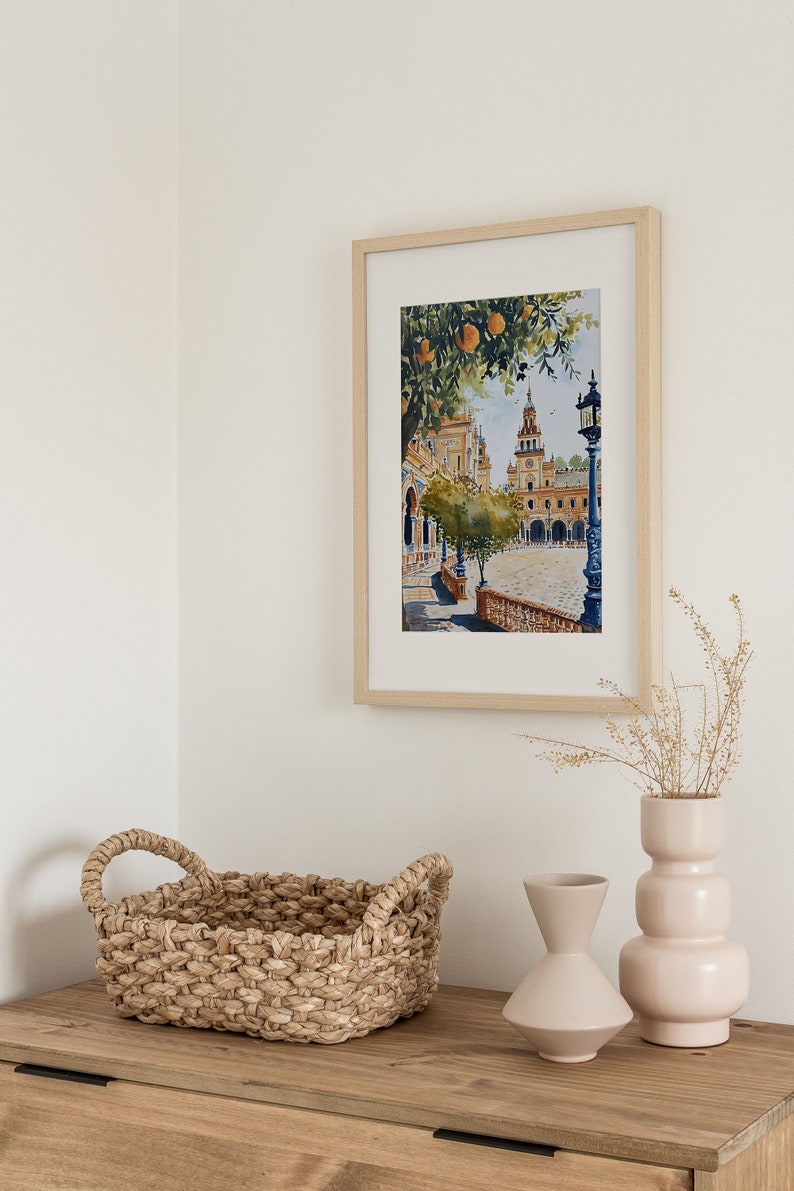May include: A framed watercolour painting of a Spanish plaza with a church in the background, a tree with orange fruit, and a street lamp. The painting is hanging on a white wall above a wooden dresser with a woven basket and two ceramic vases.