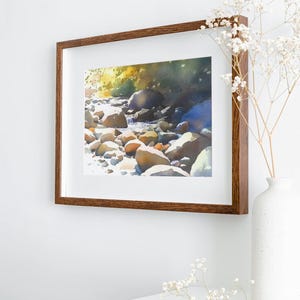 May include: A framed watercolor painting of a stream flowing through a rocky landscape. The painting is in a brown wooden frame and is hanging on a white wall. There are white flowers in a vase on a white table in the foreground.