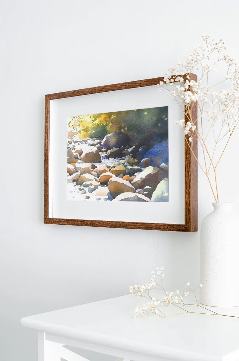 May include: A framed watercolor painting of a stream flowing through a rocky landscape. The painting is in a brown wooden frame and is hanging on a white wall. There are white flowers in a vase on a white table in the foreground.