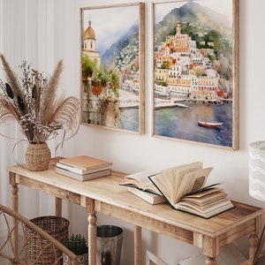 May include: Two framed watercolor paintings of a coastal town with colorful buildings on a hillside. The paintings are hung on a white wall above a wooden console table with books and a vase of dried flowers.