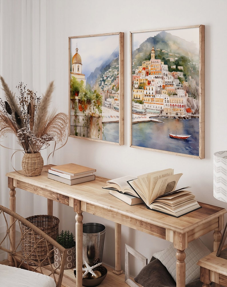 May include: Two framed watercolor paintings of a coastal town with colorful buildings on a hillside. The paintings are hung on a white wall above a wooden console table with books and a vase of dried flowers.