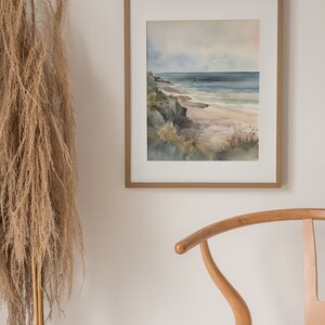 May include: A framed watercolor painting of a seascape with a rocky coastline, blue water, and a sandy beach. The painting is in a light wood frame and is hanging on a white wall.