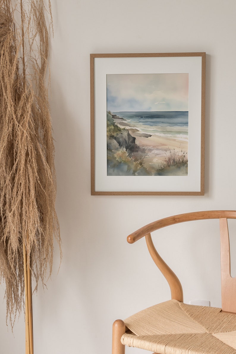 May include: A framed watercolor painting of a seascape with a rocky coastline, blue water, and a sandy beach. The painting is in a light wood frame and is hanging on a white wall.