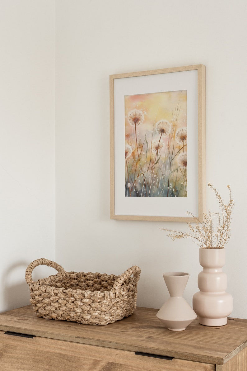 May include: A framed watercolor painting of dandelion flowers in shades of white, yellow, and brown, hanging on a white wall above a wooden dresser with a woven basket and two ceramic vases.