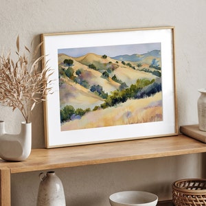 May include: A framed watercolor painting of a landscape with rolling hills and green trees. The hills are painted in shades of yellow, brown, and green. The painting is set against a white wall and a wooden shelf.