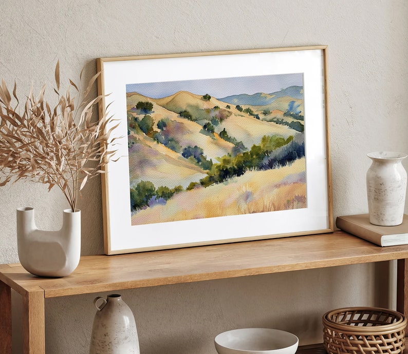 May include: A framed watercolor painting of a landscape with rolling hills and green trees. The hills are painted in shades of yellow, brown, and green. The painting is set against a white wall and a wooden shelf.