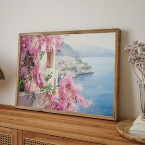 以下が含まれることがあります： 白い建物とピンクの花が咲く海岸の風景を描いた、額縁に入った水彩画。絵画は、青い海と背景の山々を望む景色を描いている。