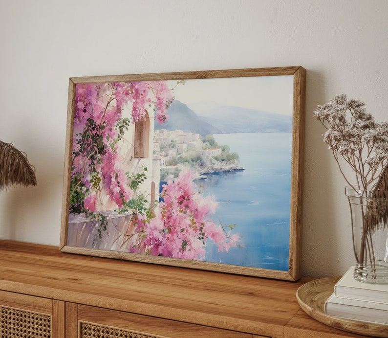 以下が含まれることがあります： 白い建物とピンクの花が咲く海岸の風景を描いた、額縁に入った水彩画。絵画は、青い海と背景の山々を望む景色を描いている。