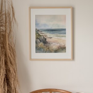May include: A framed watercolor painting of a seascape with a sandy beach, rocky cliffs, and blue ocean waves. The painting is in a light wood frame and is hanging on a white wall.
