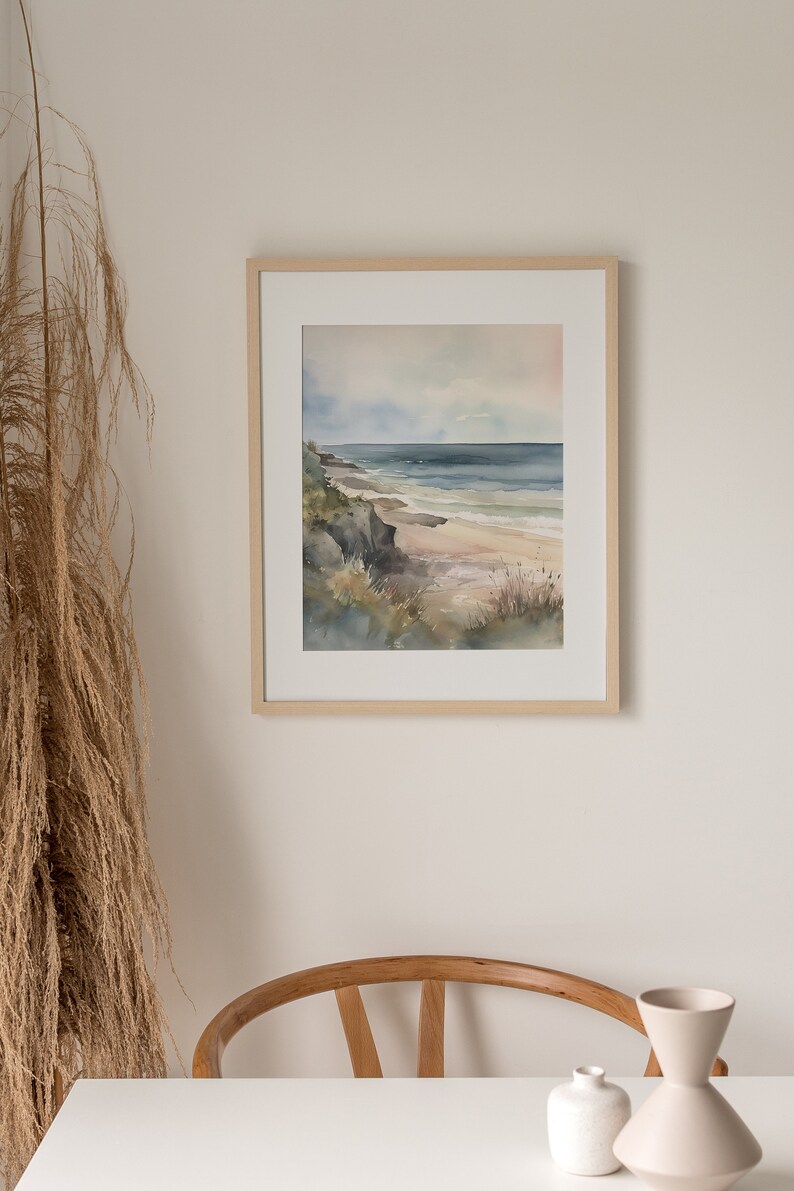 May include: A framed watercolor painting of a seascape with a sandy beach, rocky cliffs, and blue ocean waves. The painting is in a light wood frame and is hanging on a white wall.