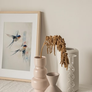 May include: A framed watercolor painting of two blue and white swallows in flight against a light blue and white background. The painting is displayed on a white table with two ceramic vases and a dried flower arrangement.