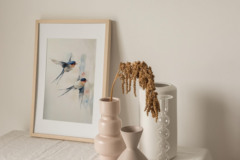 May include: A framed watercolor painting of two blue and white swallows in flight against a light blue and white background. The painting is displayed on a white table with two ceramic vases and a dried flower arrangement.