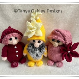 Pode incluir: Três gnomos de malha feitos à mão em tons de vermelho, amarelo e rosa. Os gnomos têm olhos de botão, cabelo de fio e usam chapéus. Um gnomo segura uma flor. O texto "© Tanya Oakley Designs" é visível.