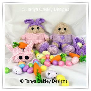 May include: Two knitted Easter egg characters, one wearing a pink dress and a purple bow, the other wearing a purple outfit, surrounded by colourful Easter eggs and carrots. © Tanya Oakley Designs