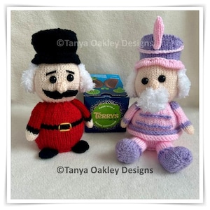 Pode incluir: Dois bonecos tricotados à mão, um com uniforme vermelho, chapéu preto e bigode, o outro com uniforme rosa e roxo com chapéu alto, posicionados ao lado de uma caixa azul de Terry's Chocolate Mint com texto branco e logotipo verde.