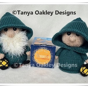May include: Two knitted gnome dolls in teal hooded jackets and hats, with beige faces and black eyes. One has a white beard, and both hold black and yellow lanterns. A box of Terry's Orange chocolate is between them. Text reads "Tanya Oakley Designs".