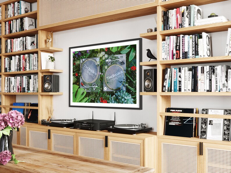 May include: Two black Technics turntables on a green and floral background. The turntables are on a shelf with a black framed picture above them. The shelf is part of a wooden bookcase with other bookshelves on either side.