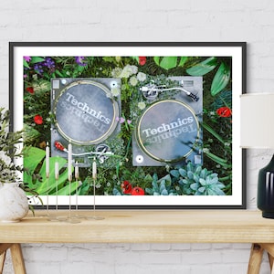 May include: A framed print of two black Technics turntables surrounded by green plants and flowers. The turntables are on a green surface. The print is framed in black and has a white mat.