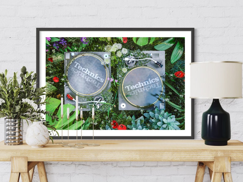 May include: A framed print of two black Technics turntables surrounded by green plants and flowers. The turntables are on a green surface. The print is framed in black and has a white mat.