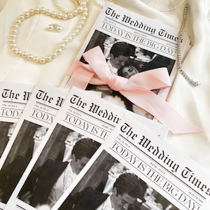 May include: A stack of wedding announcement cards with a black and white photo of a couple on the front. The cards are styled to look like a newspaper with the headline "The Wedding Times" and the text "Today is the Big Day!" The cards are tied together with a pink ribbon.
