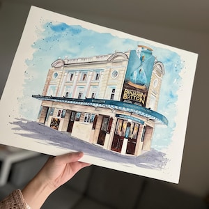 May include: Watercolour painting of a building with a sign that reads "The Curious Incident of the Dog in the Night-Time" and "Benjamin Button". The building is painted in shades of beige and brown with a blue awning. The sky is painted in shades of blue.