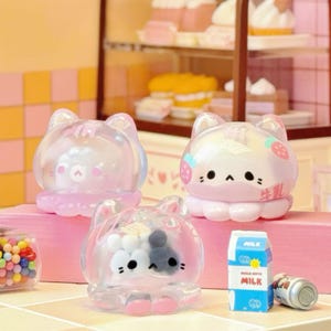 May include: Three clear, cat-shaped containers with pink accents and cartoon faces. One has a strawberry design, another has a bear face, and the third has a black and white cat face. A small carton of milk and a can are also visible.