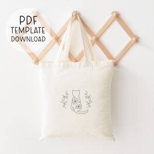 May include: A white canvas tote bag with a black line drawing of a cat with flowers on its back. The cat is sitting with its tail curled around its legs. The cat is surrounded by two sprigs of leaves.